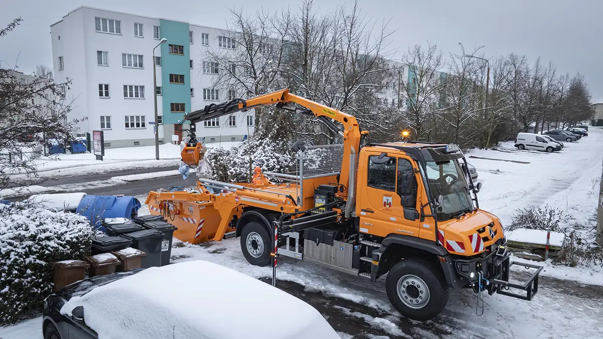 Orangefarbener Unimog Kommunal-LKW mit Ladekran und Schneepflug beim Winterdienst in einer verschneiten Wohnstrasse.