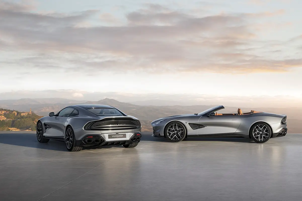 Ein silbernes Aston Martin Vanquish 25 Coupé von hinten und ein offener Vanquish 25 Volante von der Seite auf einem Aussichtspunkt.