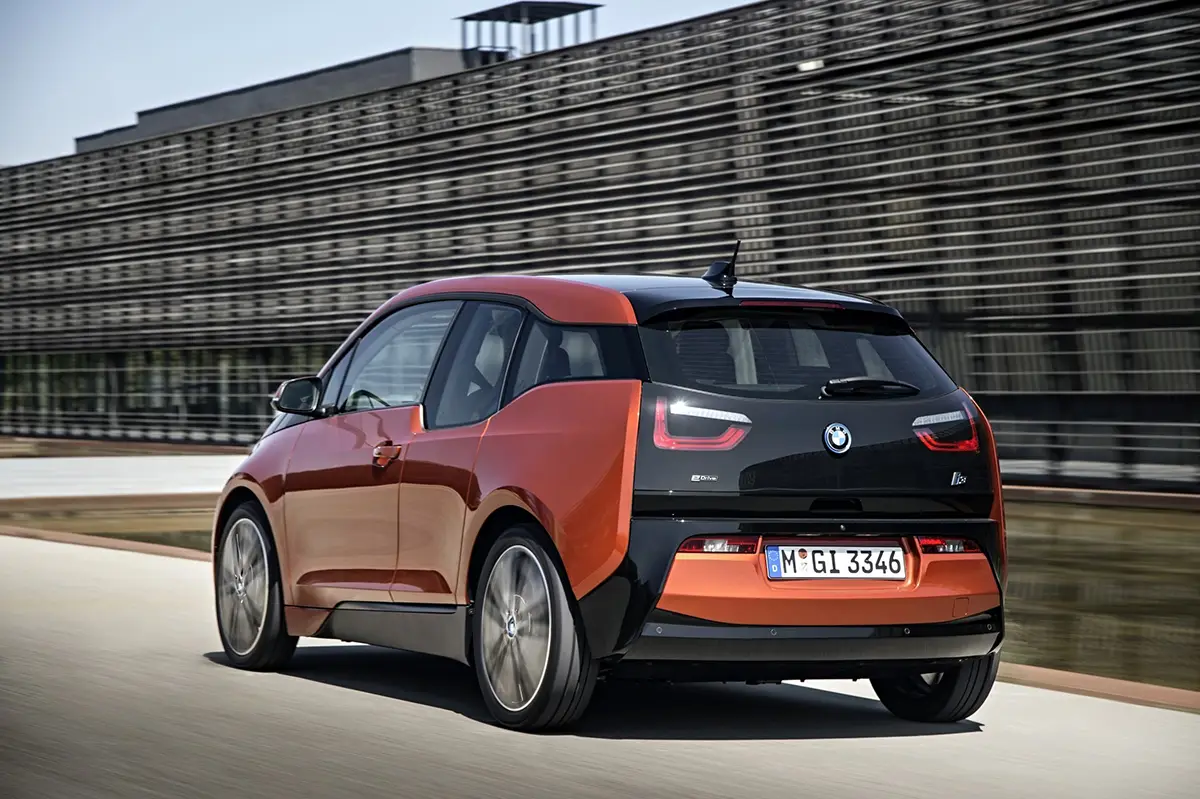 Heckseitenansicht eines orange-schwarzen BMW i3 Elektro-Kleinwagens vor einem modernen Gebäude mit Holzlamellen-Fassade.