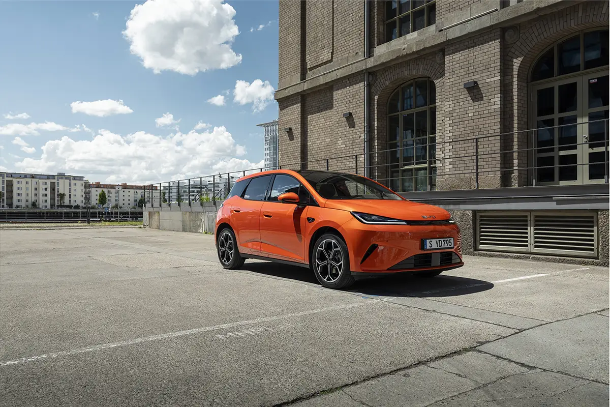 Ein kompakter, orangefarbener BYD Dolphin parkiert auf einem urbanen Betonplatz direkt vor der Fassade eines historischen Fabrikgebäudes aus Backstein.
