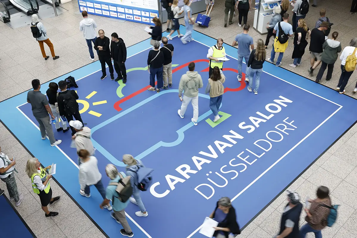 Besucherinnen und Besucher laufen in der Messe Düsseldorf über einen grossen, blauen Messeteppich mit der weissen Aufschrift Caravan Salon Düsseldorf.