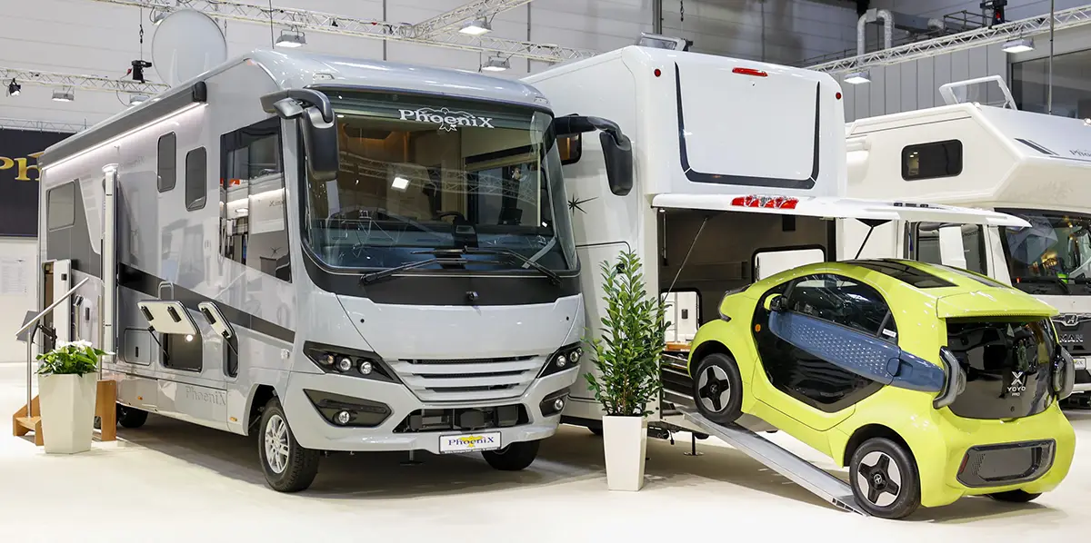 Ein grosses, silbernes Luxus-Reisemobil auf der Messe mit geöffneter Heckgarage, aus der ein kleines gelbes Elektroauto über eine ausgefahrene Laderampe herausrollt.