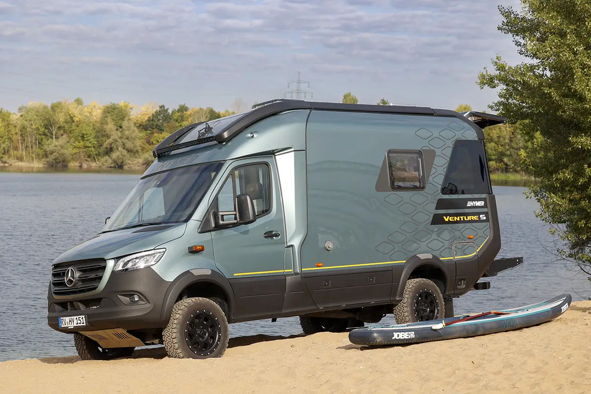 Ein geländegängiges Allrad-Reisemobil (Crossover-Modell auf Mercedes-Benz-Basis) in Graugrün parkt auf einer Sandfläche direkt an einem Seeufer neben einem abgelegten Kajak.