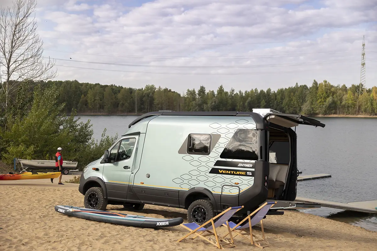 Heckansicht eines kompakten Offroad-Camper-Vans am Sandstrand eines Sees mit weit geöffneten Heckklappen und montierter Einstiegsrampe.