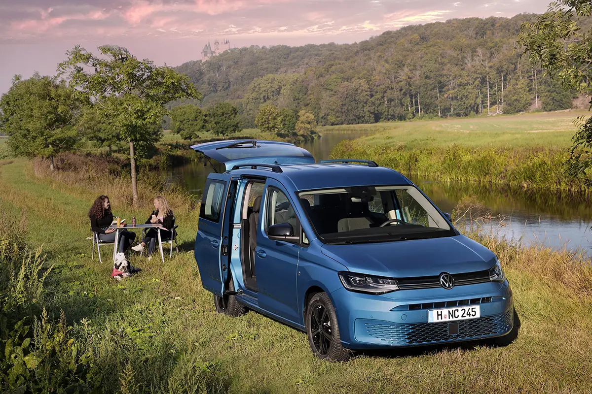 Ein blauer VW Caddy California steht mit geöffneter Heckklappe auf einer Wiese an einem Flussufer, im Hintergrund entspannen zwei Personen auf Campingstühlen.