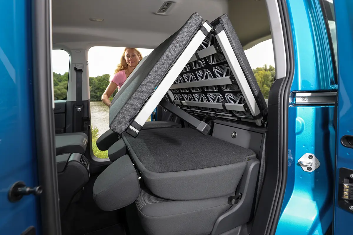 Blick durch die geöffnete Schiebetür eines blauen VW Caddy, bei dem der innovative, klappbare Lattenrost des Schlafsystems nach vorne entfaltet wird.