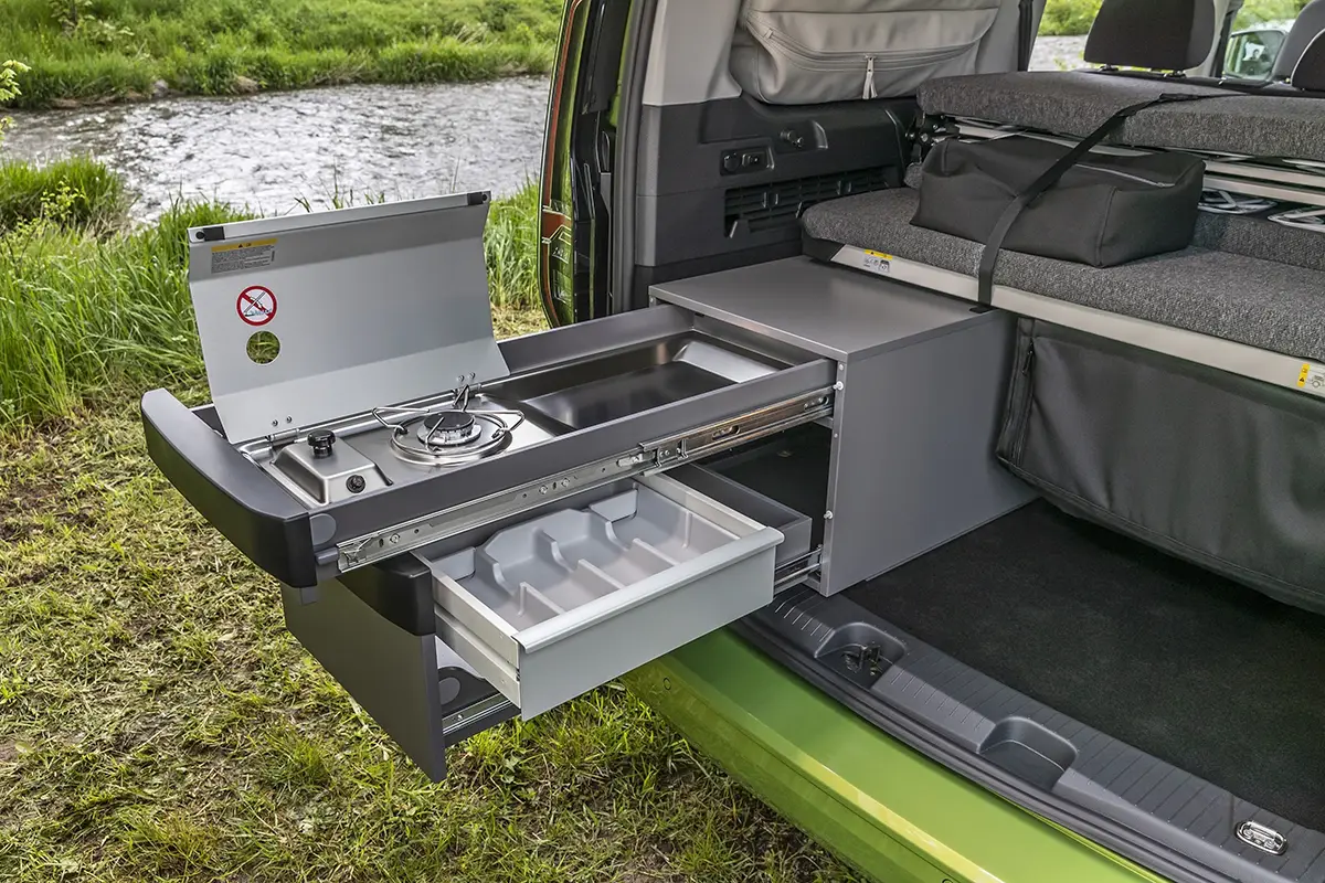 Detailaufnahme der optionalen, ausziehbaren Miniküche im Heck des VW Caddy California mit aufgeklapptem Einflammen-Gasherd am Flussufer.