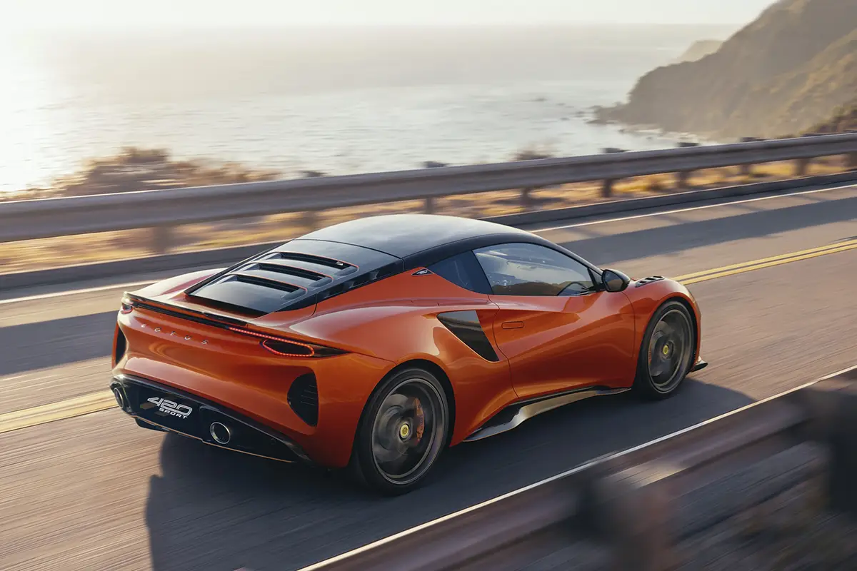 Heckseitenansicht des orangefarbenen Lotus Emira 420 Sport bei dynamischer Fahrt auf einer asphaltierten Küstenstrasse.
