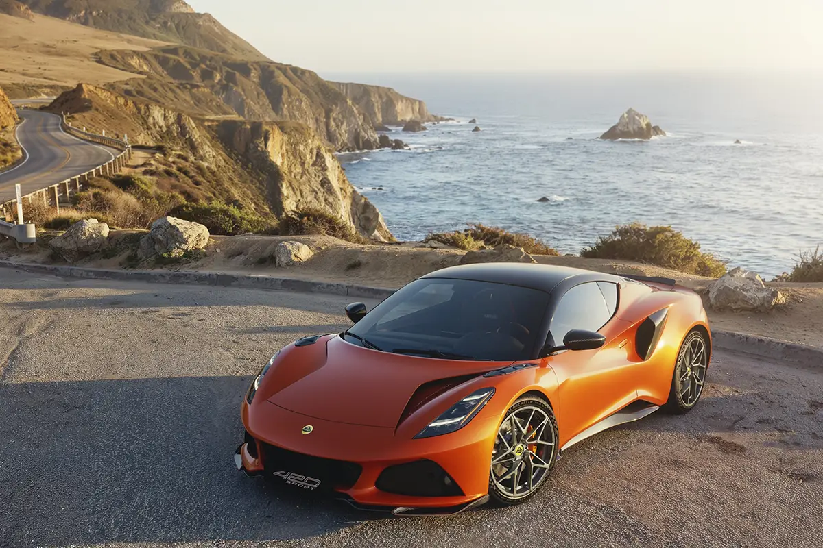 Ein orangefarbener Lotus Emira 420 Sport Mittelmotor-Sportwagen parkiert auf einem Aussichtspunkt an einer Küstenstrasse vor dem Ozean.