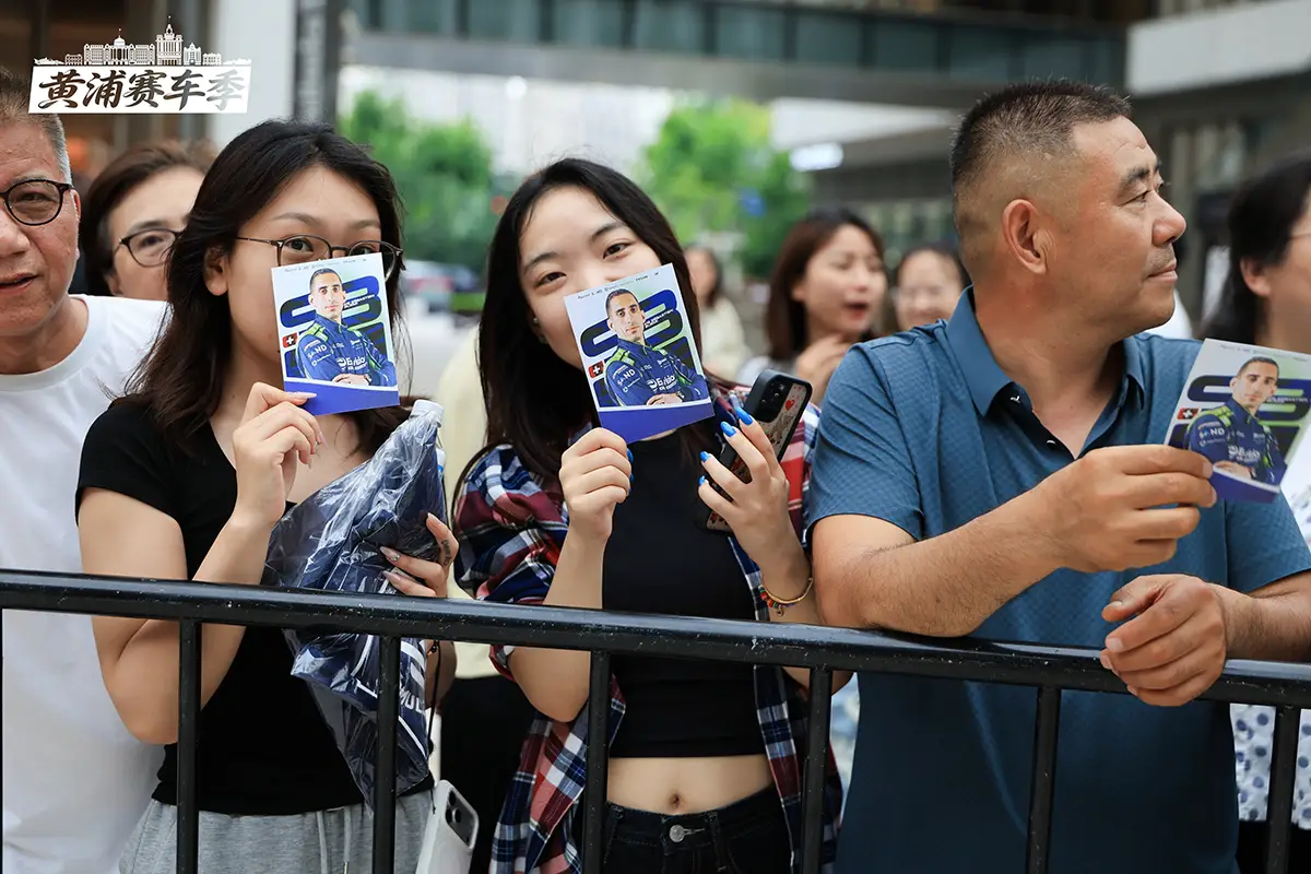 Zwei junge chinesische Frauen an einem Sicherheitszaun halten lächelnd Autogrammkarten des Schweizer Rennfahrers Sébastien Buemi beim Envision Racing Roadshow-Event in Shanghai hoch.