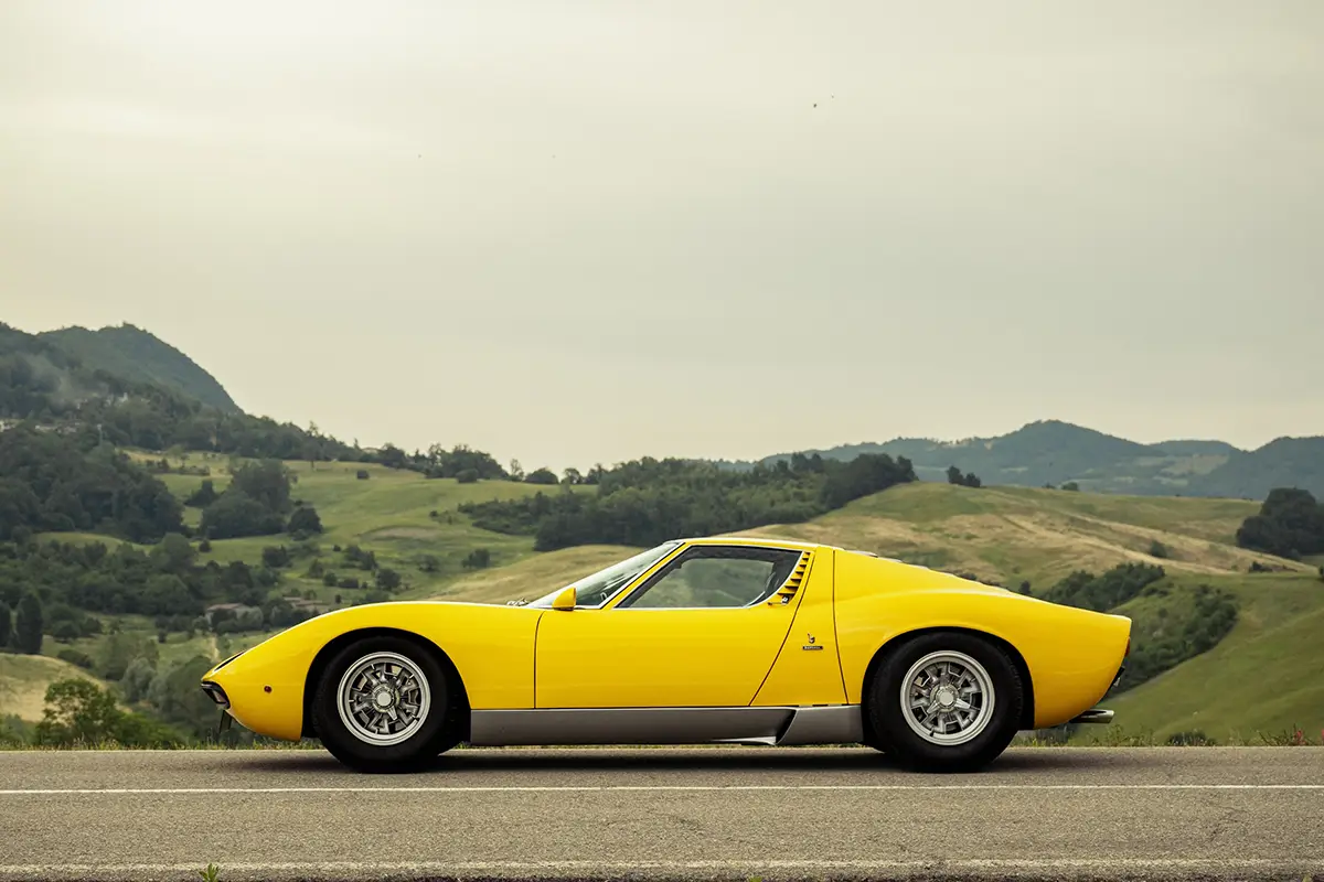Profilansicht der flachen, fliessenden Linienführung des gelben Lamborghini Miura Klassikers auf einer Landstrasse.