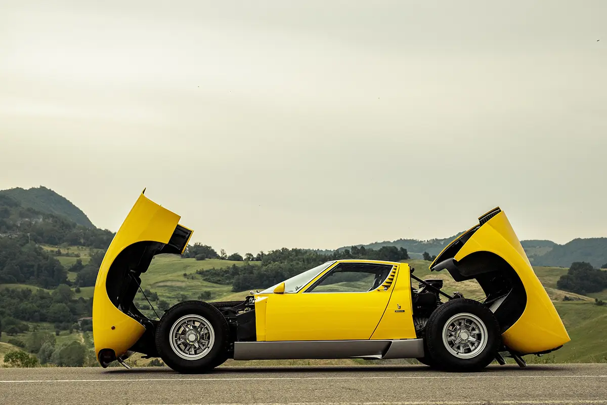 Seitenansicht des gelben Lamborghini Miura mit komplett nach vorne geklappter Fronthaube und nach hinten geklappter Motorabdeckung, die den Blick auf das Chassis freigibt.