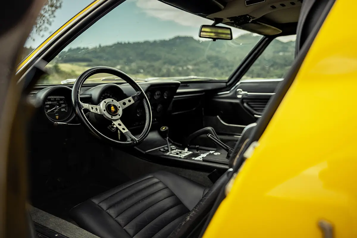 Blick in das enge, puristische Cockpit des Lamborghini Miura mit Blick auf die analogen Rundinstrumente, die offene Schaltkulisse und die schwarzen Ledersitze.