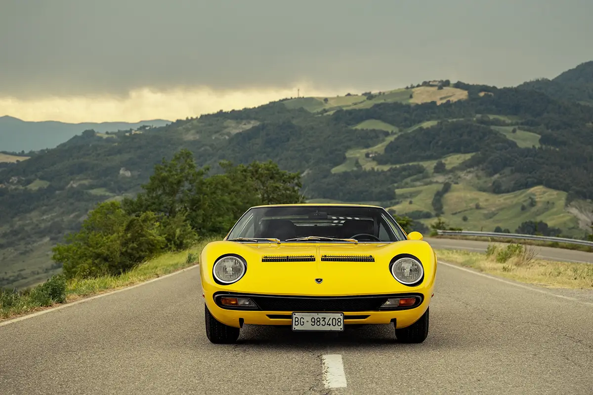Frontalansicht eines klassischen gelben Lamborghini Miura Supersportwagens auf einer asphaltierten Landstrasse vor einer hügeligen Landschaft.