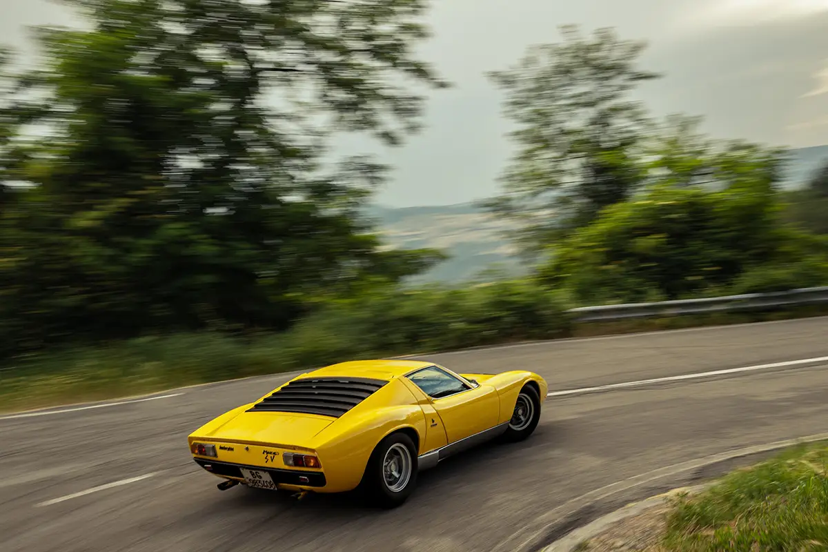 Heckseitenansicht eines gelben Lamborghini Miura in dynamischer Fahrt auf einer kurvigen Landstrasse.