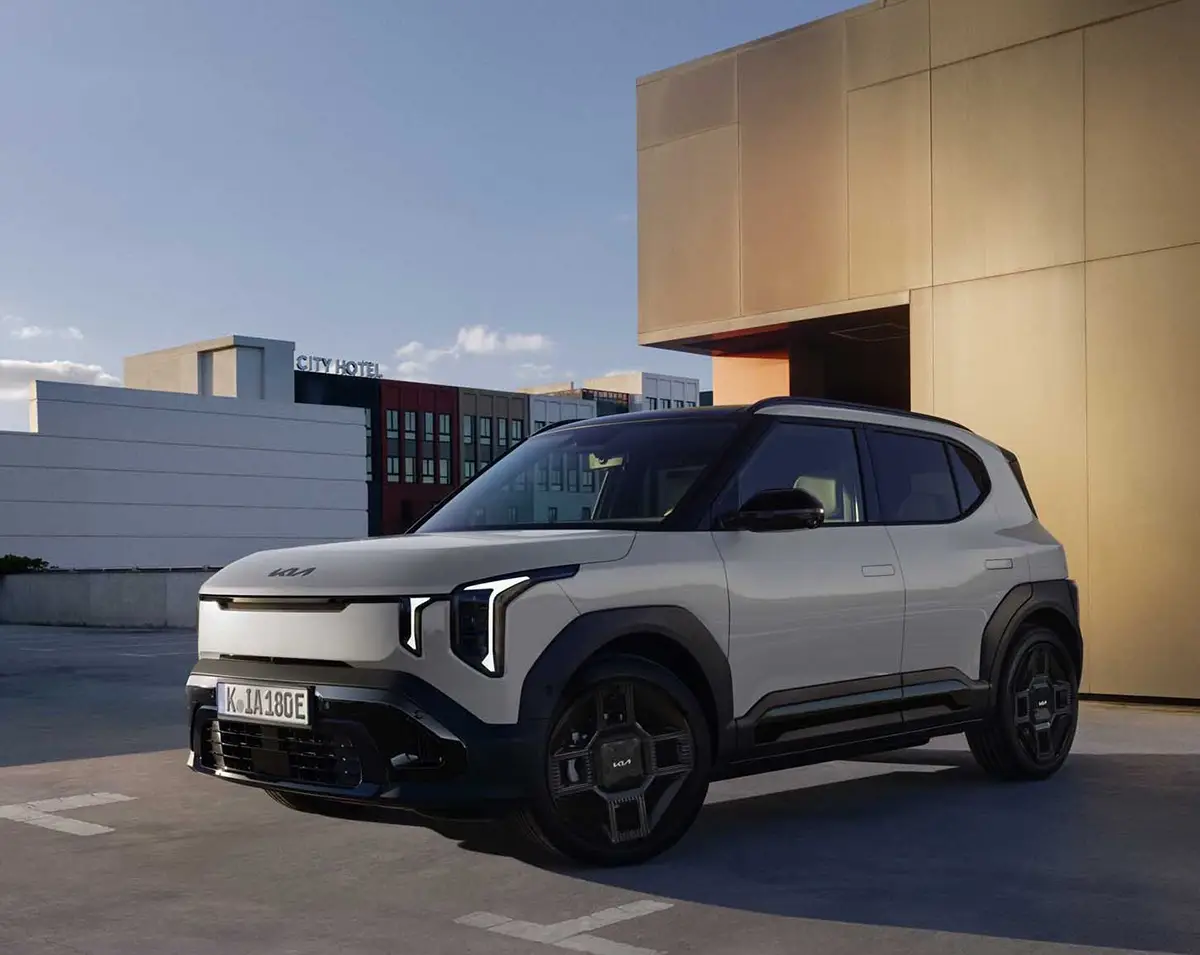 Ein hellgrauer, kantiger Kia EV2 Elektro-Crossover steht auf dem Parkdeck eines modernen Gebäudes im Sonnenlicht.