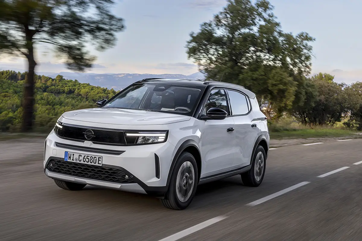 Ein weisser Opel Frontera Electric fährt dynamisch auf einer Landstrasse an Bäumen vorbei.