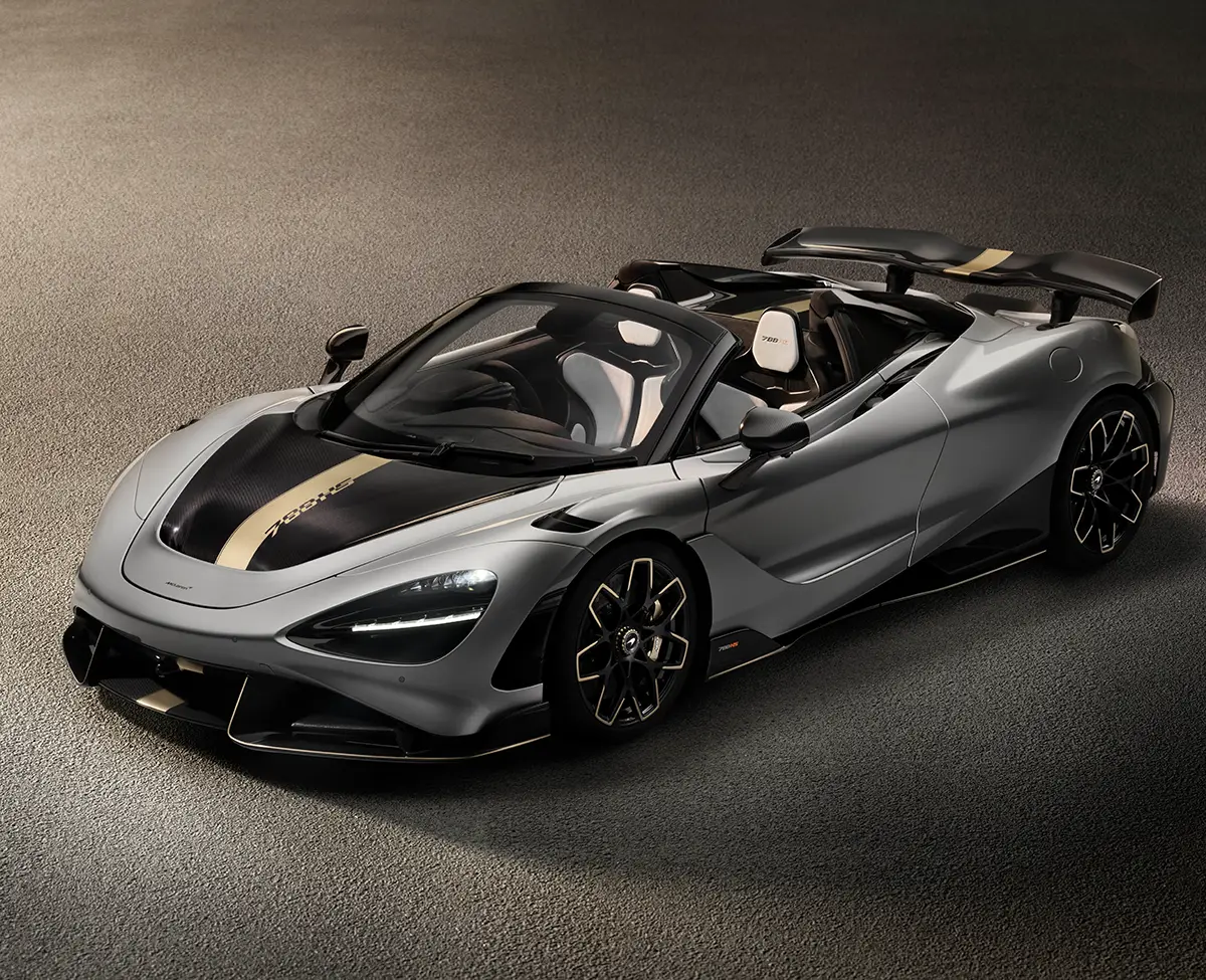 Silver McLaren 720S Spider convertible with a gold racing stripe, shown from the front-left on a textured asphalt surface. The car features a large rear wing and tan interior seats visible through the open top.