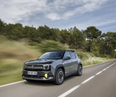 Blue-gray Renault SUV driving on a rural two-lane road with trees and blue sky ahead of it