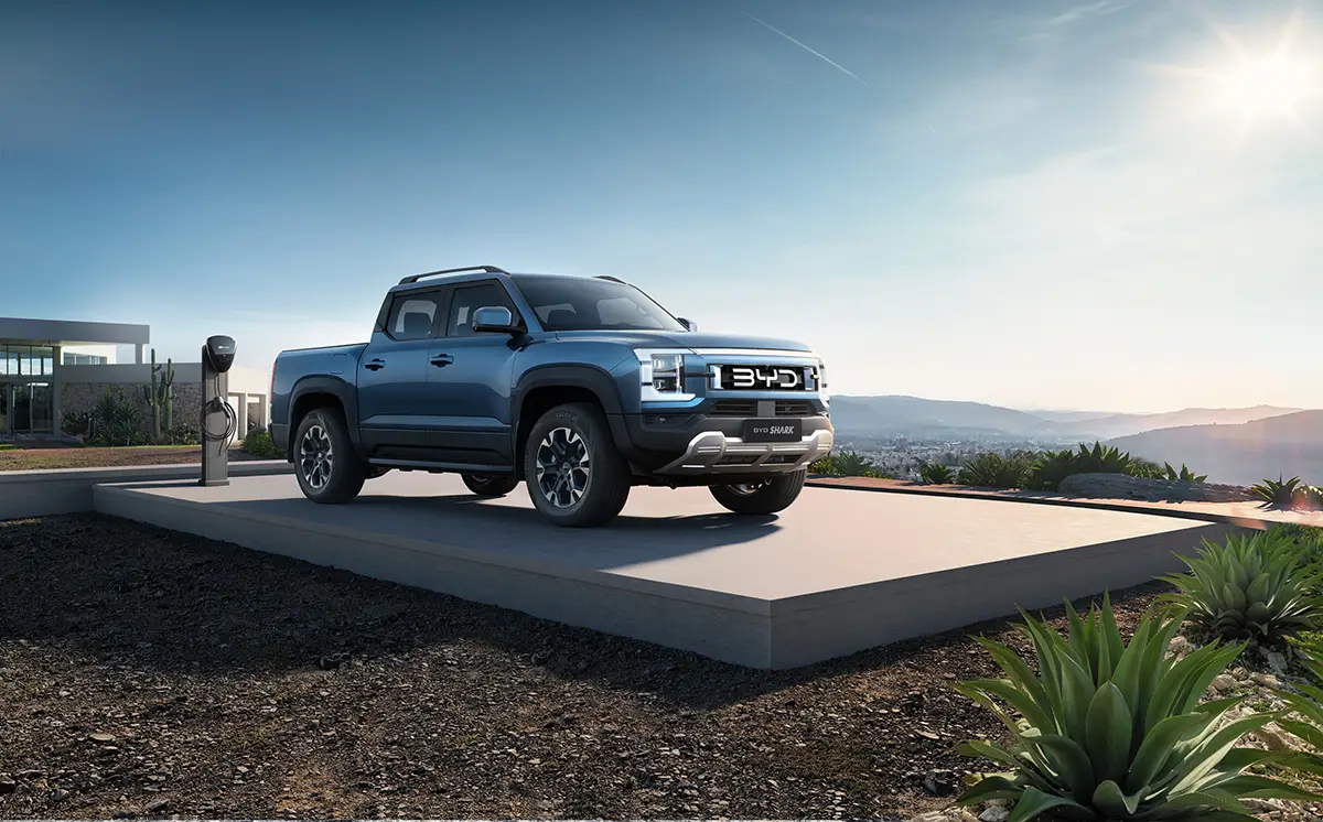 Der neue, dunkelblaue BYD Shark Plug-in-Hybrid-Pickup steht im Freien auf einer Betonplattform neben einer Ladestation vor einer Hügellandschaft bei Sonnenschein.