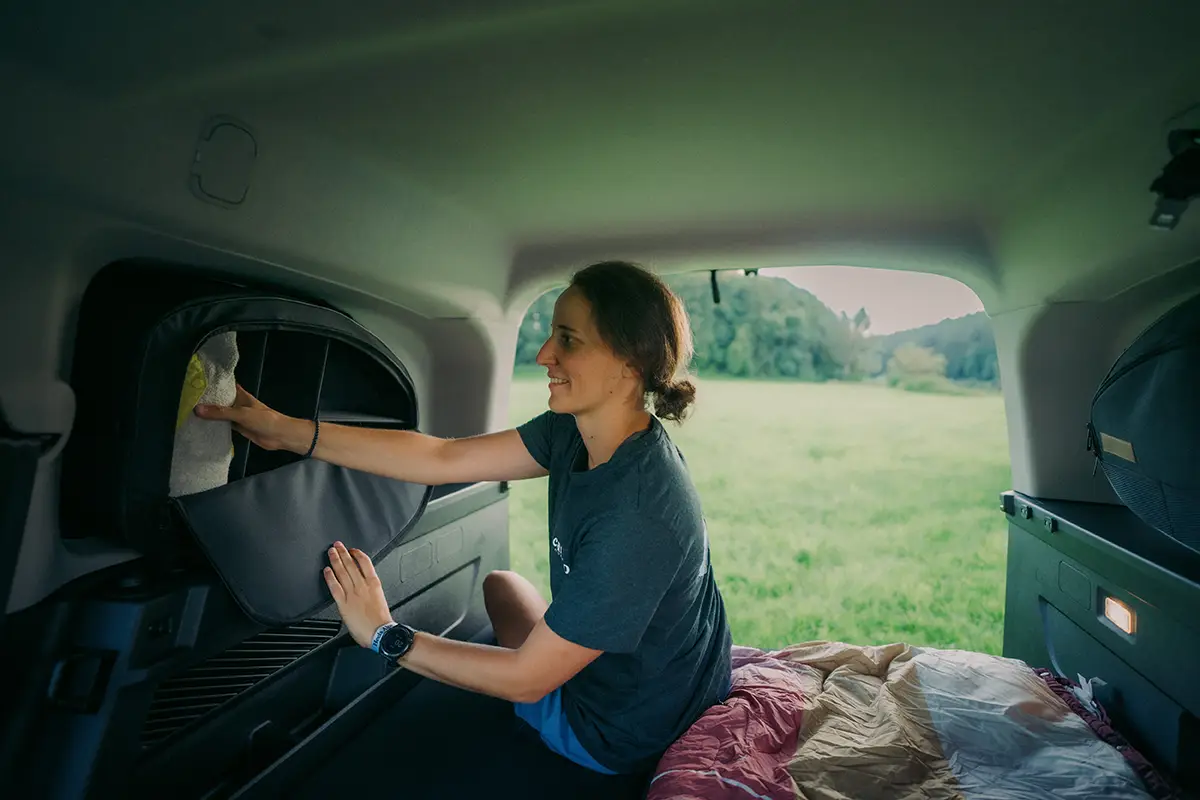 Innenansicht eines kompakten Campers, bei dem eine Camperin im Fond auf dem Schlafsystem sitzt und Gepäck in den seitlich montierten Fenstertaschen verstaut.