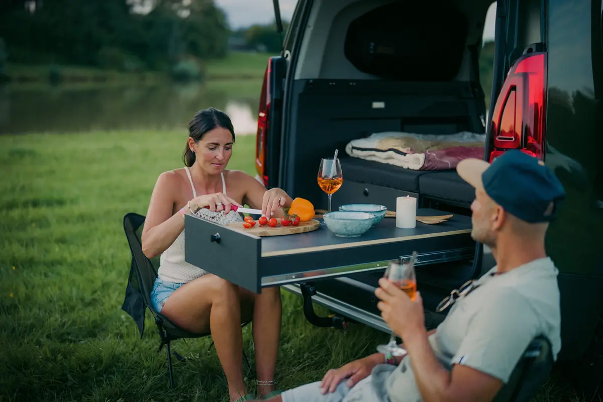 Zwei Camper sitzen im Freien auf Campingstühlen an einem ausfahrbaren Heckküchen-Tisch eines Micro-Campers und geniessen ein Abendessen an einem See.