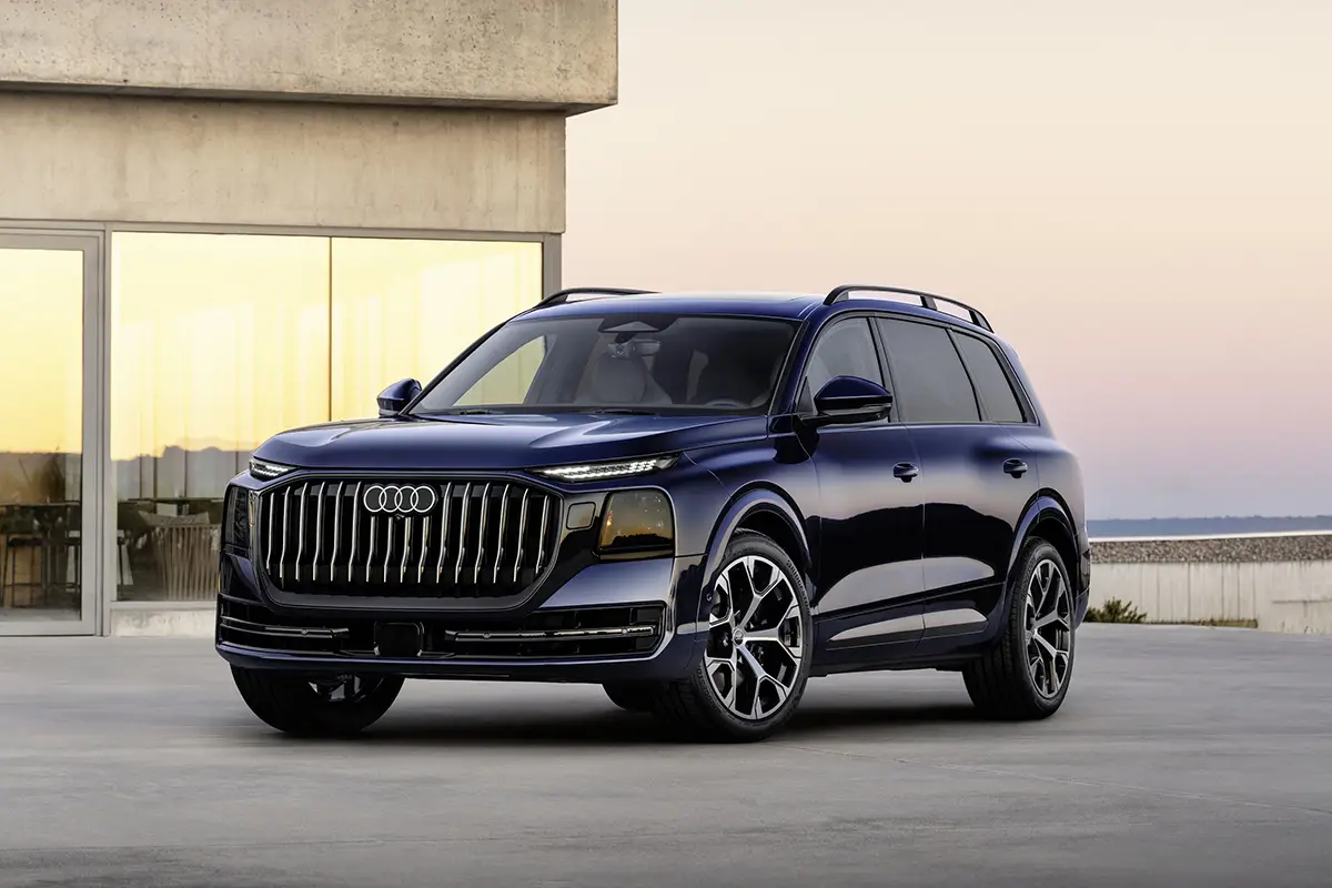 Schrägansicht von vorne auf den dunkelblauen Audi Q9 XXL-SUV mit vertikalen Lamellen im Singleframe-Kühlergrill vor moderner Architektur.