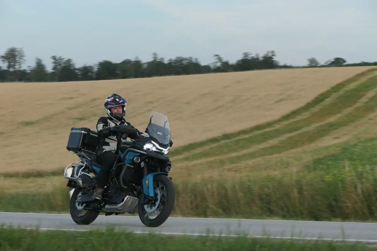 Seitliche Fahraufnahme der CF Moto 800 MT-ES mit Gepäcksystem auf einer asphaltierten Landstrasse vor Getreidefeldern.