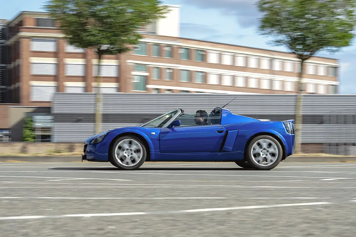 Seitenansicht des offenen blauen Opel Speedster in dynamischer Fahrt vor Backsteingebäuden.