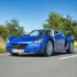 Blauer Opel Speedster fährt mit geöffnetem Stoffverdeck auf einer Landstrasse im Sonnenlicht.