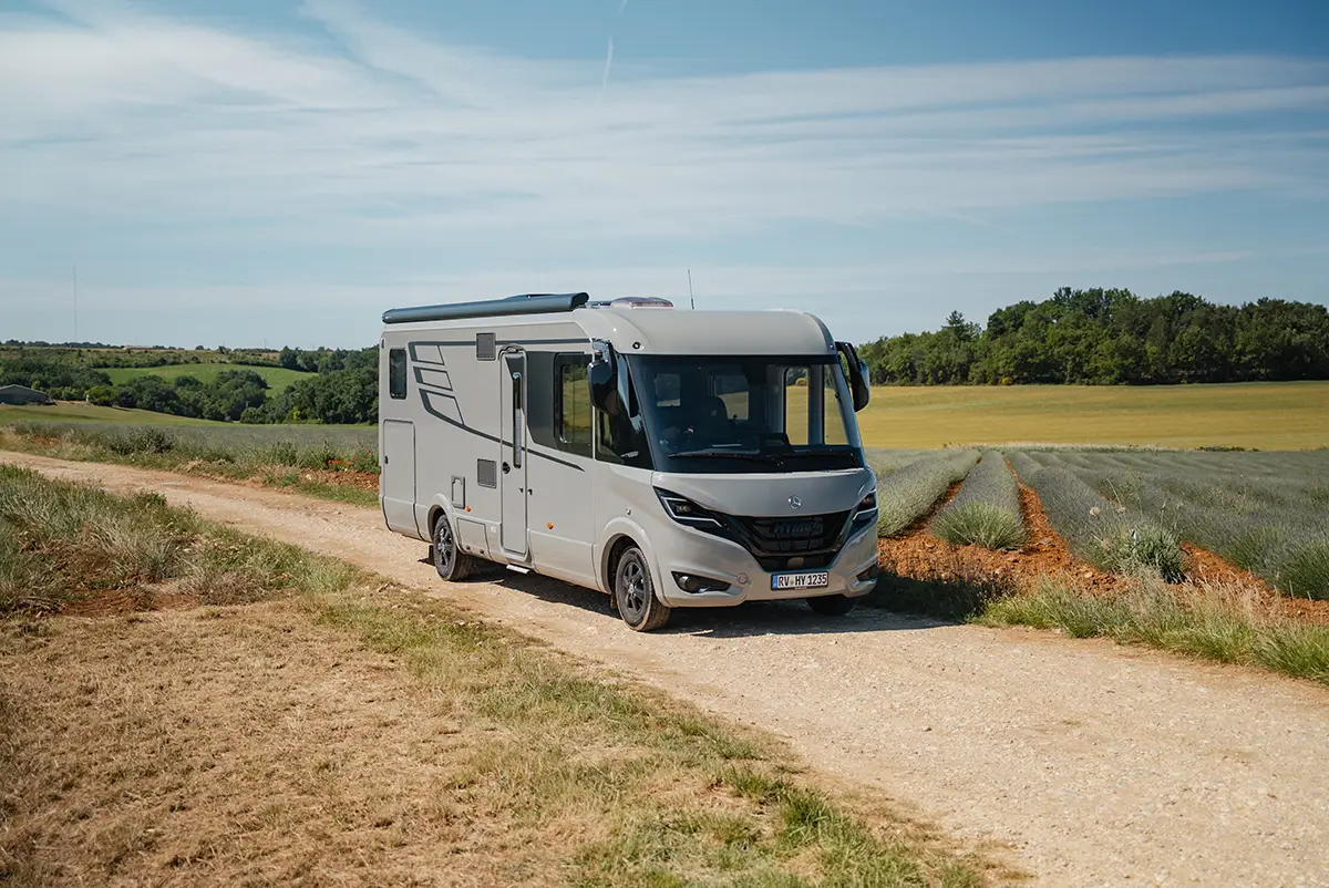 Der vollintegrierte Hymer B-Klasse Master-Line in hellem Premium-Grau fährt auf einem unbefestigten Feldweg an einem Getreidefeld vorbei.