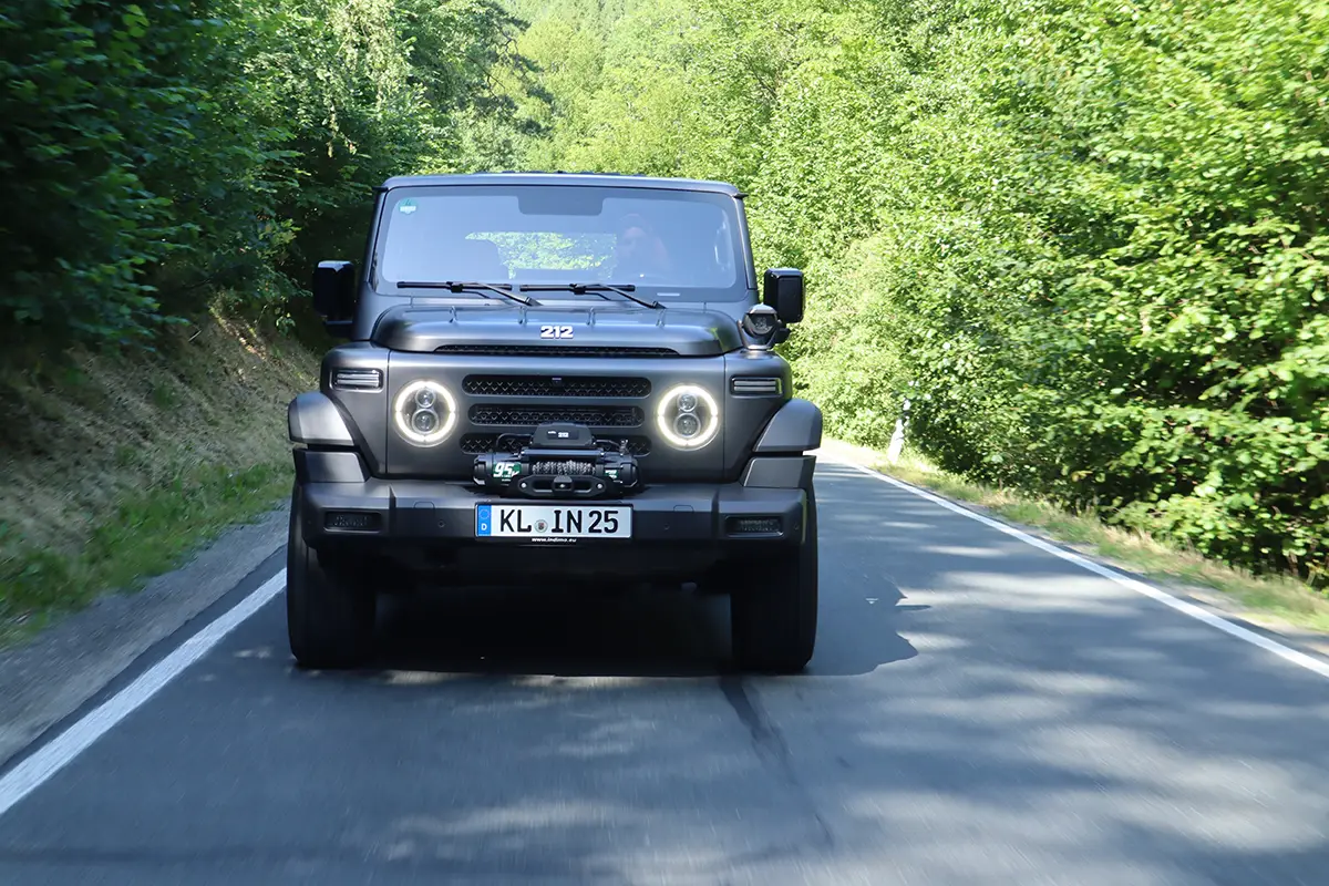 Frontalansicht des kantigen, dunkelgrauen BAW 212 Geländewagens mit leuchtenden LED-Rundscheinwerfern auf einer schattigen Landstrasse.