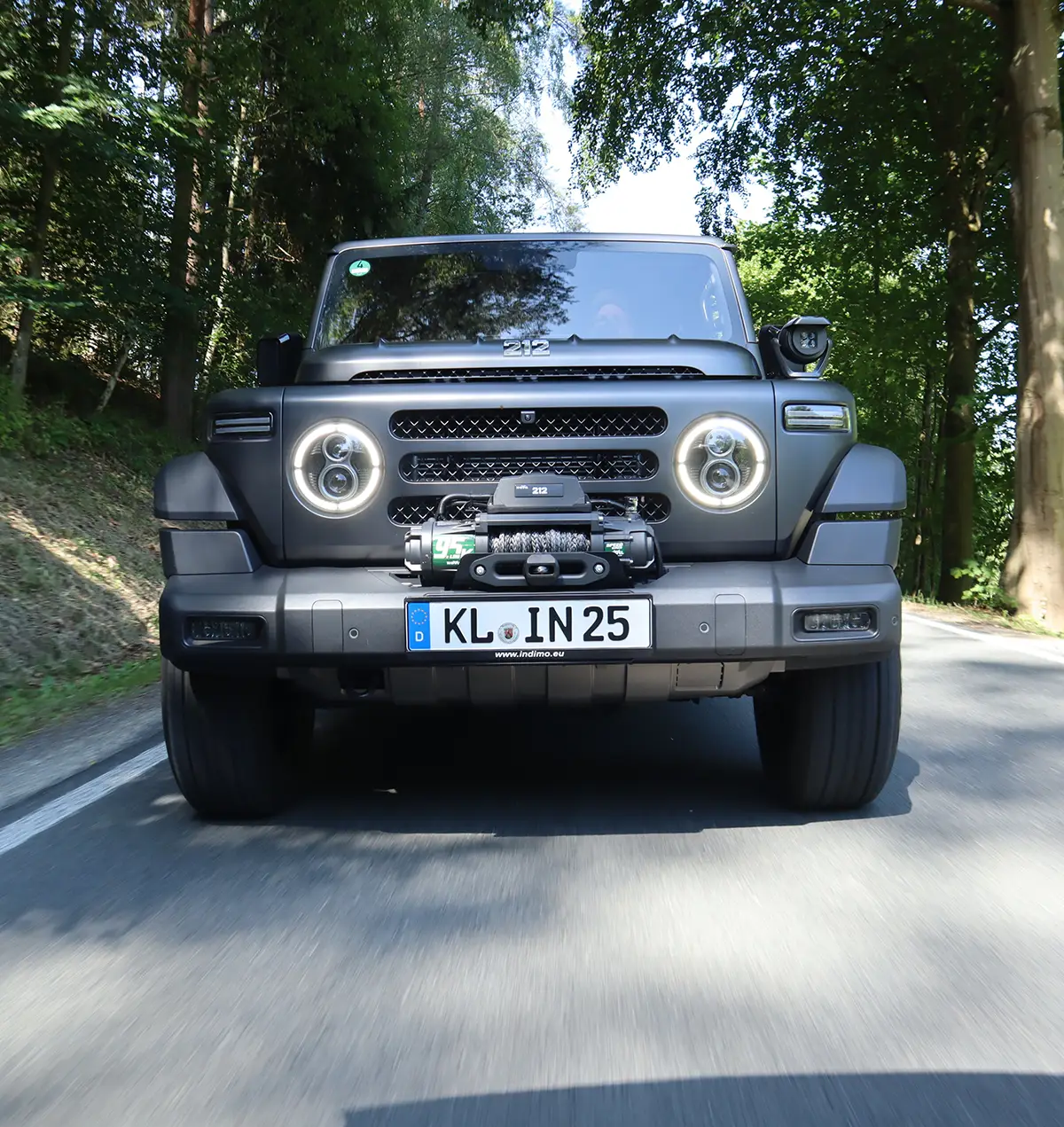 Der BAW 212 nähert sich frontal auf einer asphaltierten Waldstrasse mit eingeschalteten runden LED-Tagfahrleuchten.