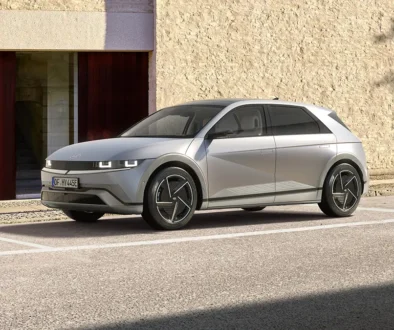 Silver futuristic hatchback parked in a parking lot beside a beige stone wall and wooden doors.