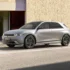 Silver futuristic hatchback parked in a parking lot beside a beige stone wall and wooden doors.