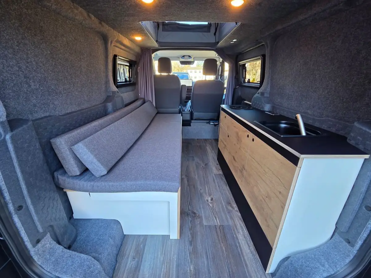 Innenraum des Alpincamper Kia PV5 mit langer grauer Schlafcouch auf der linken Seite und Holz-Küchenzeile rechts.