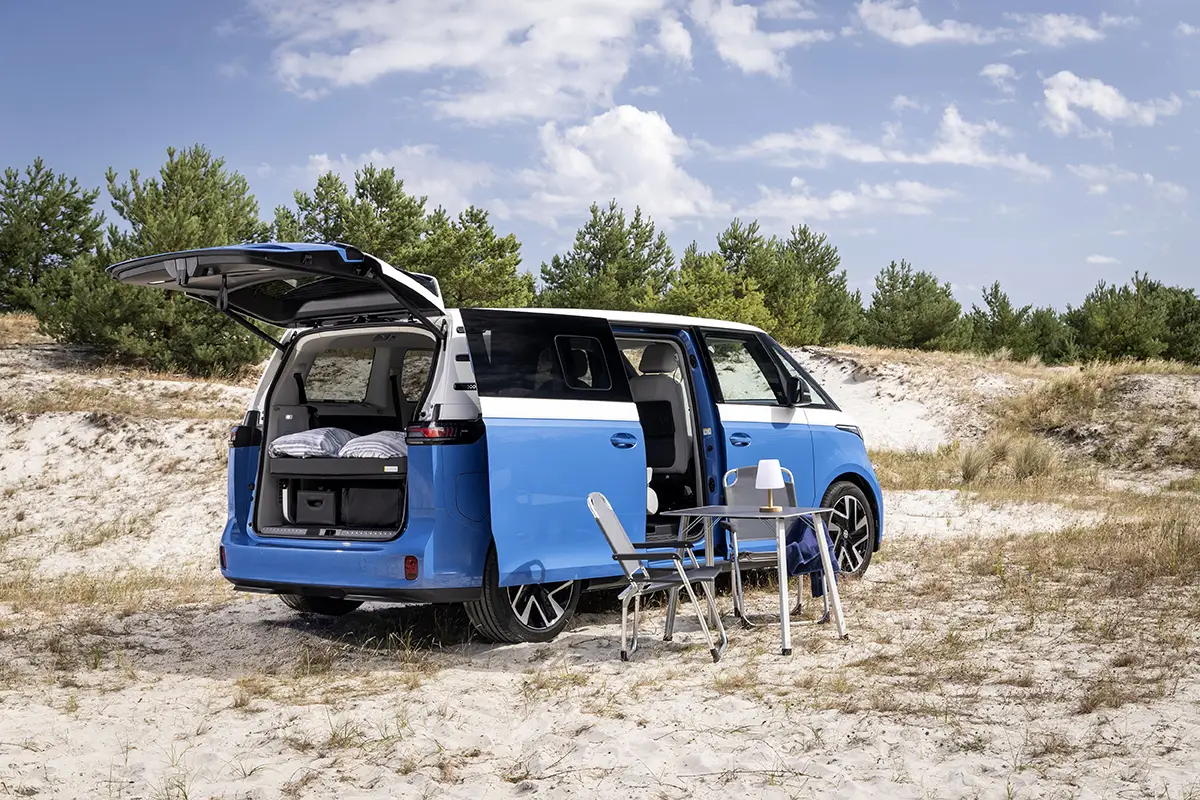 Zweifarbiger VW ID.California Cruise Elektro-Camper mit geöffneter Heckklappe und Campingmöbeln im Sand.