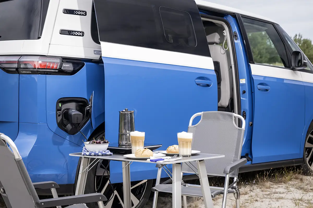 Camping-Setup mit Tisch, Klappstühlen und Kaffeegläsern neben der offenen Schiebetür des elektrischen VW ID.California Cruise.