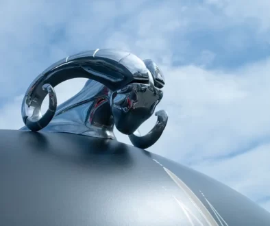 Detailaufnahme der verchromten Dodge Widder Kühlerfigur mit geschwungenen Hörnern auf einer mattschwarzen Oldtimer-Motorhaube gegen blauen Himmel