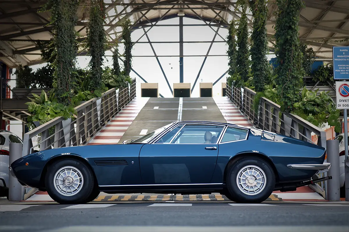 1972er Maserati Ghibli SS Coupé im Profil mit verchromten Fensterrahmen und klassischen Alufelgen vor einer Industrieüberdachung. Foto: Maserati