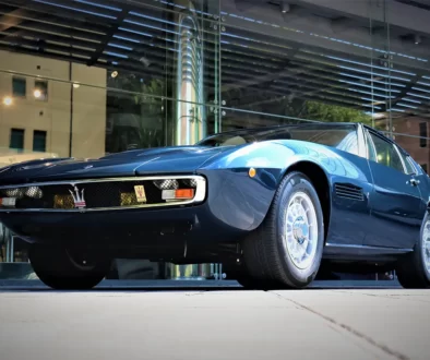 Frontansicht des blauen 1972er Maserati Ghibli SS Coupé mit flacher Haube und markantem Dreizack-Kühlergrill vor moderner Glasfassade. Foto: Maserati